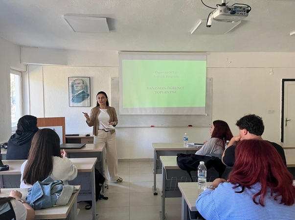 Dalaman MYO’da bir sınıfta düzenlenen danışman–öğrenci toplantısı. Önde bir öğretim elemanı kürsü yanında ayakta durarak sunum yapıyor. Arkadaki projeksiyon perdesinde ‘Dalaman MYO Lojistik Programı Danışman-Öğrenci Toplantısı’ başlığı görünüyor. Sınıfta sıralarda oturan öğrenciler sunumu dikkatle dinliyor. Duvarlarda Atatürk portresi ve büyük pencerelerden gün ışığı yansıyor.